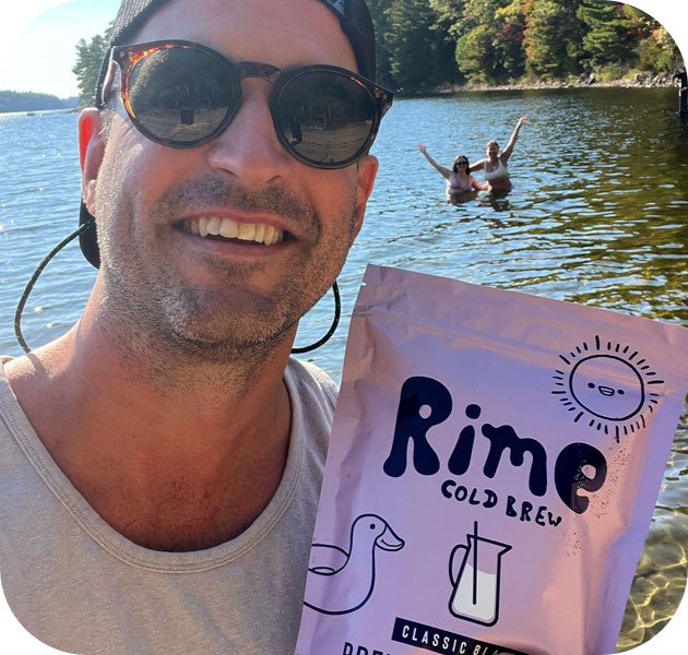 Man holding a Rime Cold Brew package with people in the background by a lake